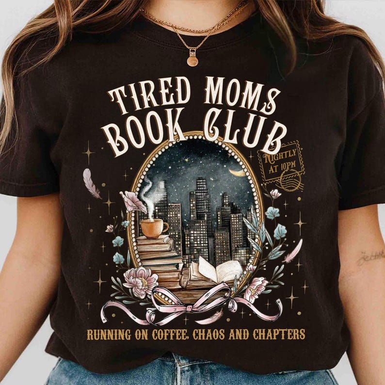 May include: Dark brown t-shirt with the text "TIRED MOMS BOOK CLUB" above an oval illustration of a cityscape, coffee cup, and books. The text "RUNNING ON COFFEE, CHAOS AND CHAPTERS" is below the illustration.