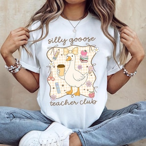 May include: White t-shirt with a cartoon goose design. The goose is holding a coffee cup and wearing a bow. The shirt has the words "silly goooose teacher club" in a decorative frame with school-themed illustrations.