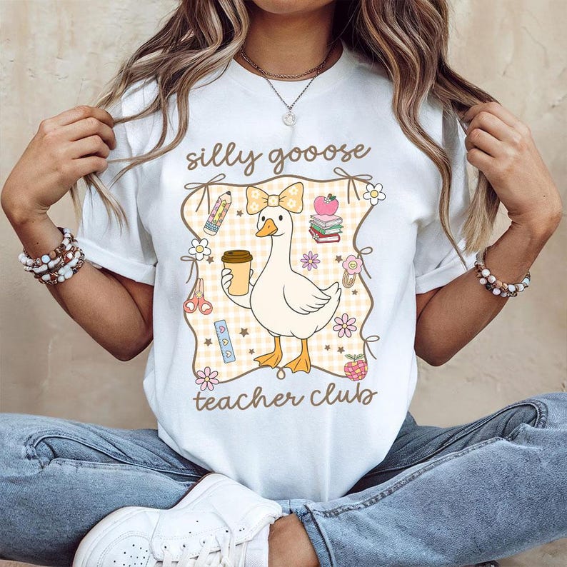 May include: White t-shirt with a cartoon goose design. The goose is holding a coffee cup and wearing a bow. The shirt has the words "silly goooose teacher club" in a decorative frame with school-themed illustrations.