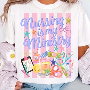 May include: White t-shirt with the text "Nursing is my Ministry" in blue script, decorated with stars and medical-themed illustrations. Pink and white striped background. Includes a stethoscope, scissors, and a smiley face.