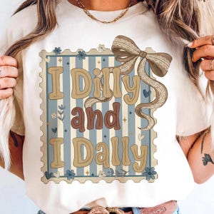 May include: Cream-colored t-shirt featuring the phrase "I Dilly and I Dally" in a vintage-style design. The text is set within a blue and white striped frame, adorned with floral and bow accents. The bow has a lace trim.