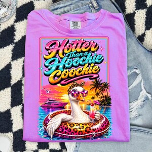 May include: Lilac Comfort Colors t-shirt with a graphic of a goose wearing sunglasses and a leopard print headband, floating in a pool with a cocktail. The text reads "Hotter Than a Hoochie Coochie."
