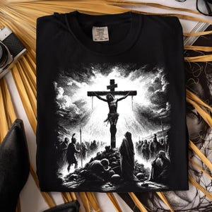 May include: Black t-shirt featuring a detailed black and white illustration of Jesus on the cross, with figures at the base and a dramatic sky. The shirt has a Comfort Colors tag.