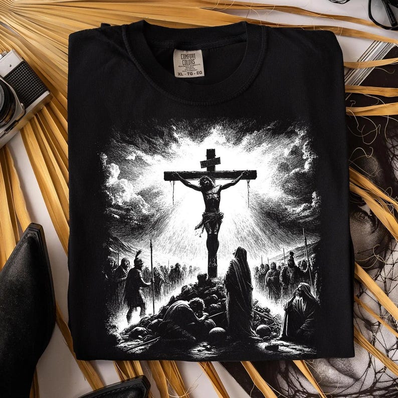 May include: Black t-shirt featuring a detailed black and white illustration of Jesus on the cross, with figures at the base and a dramatic sky. The shirt has a Comfort Colors tag.