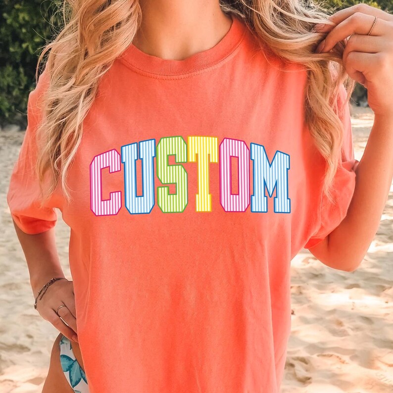 May include: Coral-colored t-shirt with the word "CUSTOM" in a colorful, block letter design. The letters are outlined in a dark blue, with a striped pattern in the letters. The shirt has a crew neck and short sleeves.