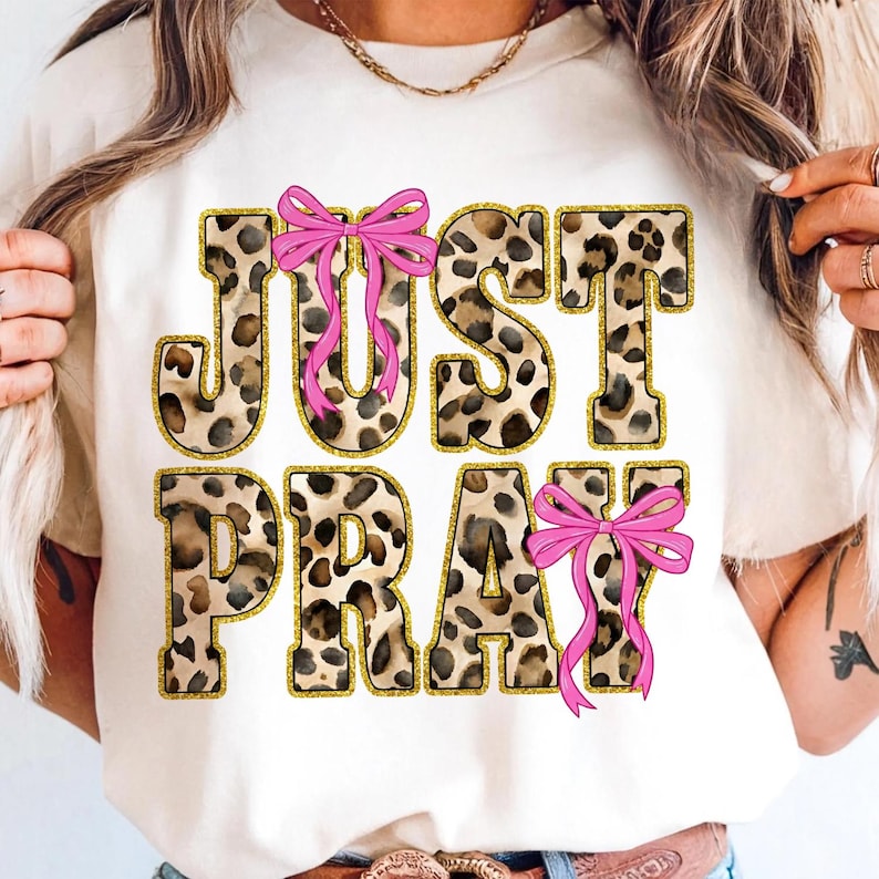 May include: Cream-colored t-shirt with the words "JUST PRAY" in a leopard print design outlined in gold glitter. Pink bows are placed above the "U" and below the "Y".