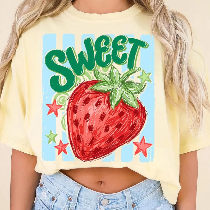May include: Yellow t-shirt with a large graphic of a red strawberry and the word "SWEET" in green. The strawberry has green leaves and is surrounded by red and green stars. The background is light blue with vertical stripes.