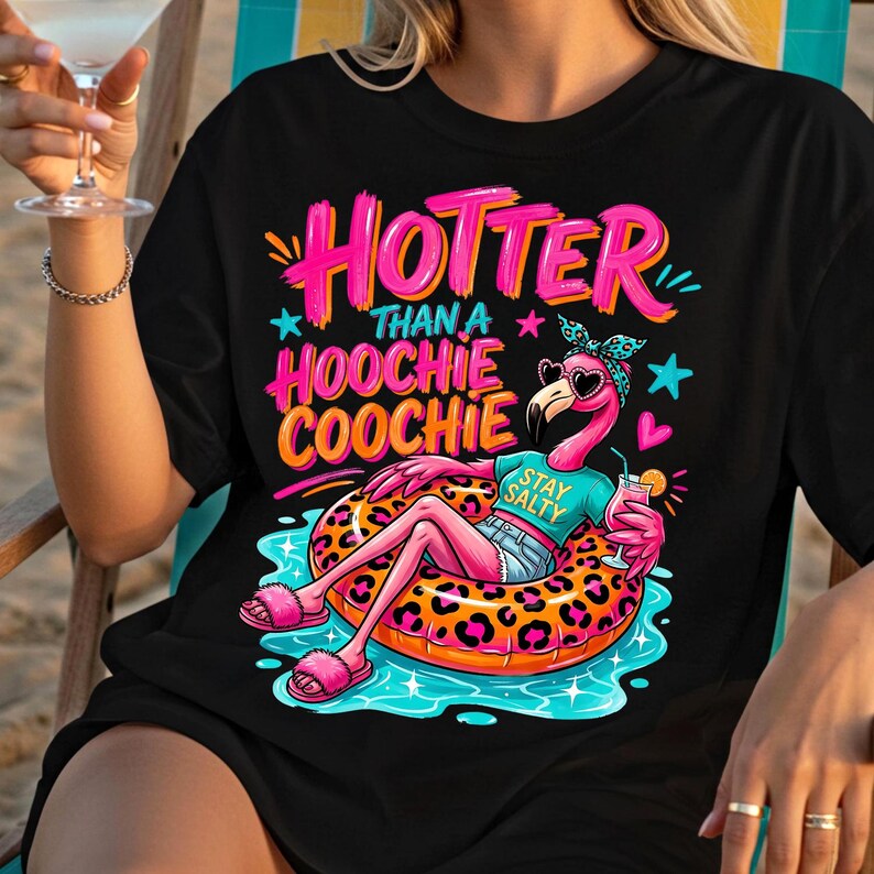 K&ouml;nnte beinhalten: Schwarzes T-Shirt mit einem Flamingo auf einem Schwimmring. Der Text lautet "Hottie Than a Hoochie Coochie" in Pink und Orange. Der Flamingo tr&auml;gt eine Sonnenbrille und ein Stirnband und h&auml;lt einen Cocktail.