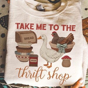 以下が含まれることがあります： クリーム色のスウェットシャツ。ガチョウ、バスケットに入った鶏、パン、ボウル、そして「TAKE ME TO THE thrift shop」の文字が描かれたグラフィックデザイン。デザインにはキノコとシナモンの容器が含まれています。