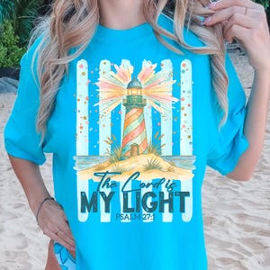 Pu&ograve; includere: Maglietta turchese con un design a faro colorato. Il faro ha strisce rosse, arancioni e verdi. Il testo recita "The Lord is My Light" e Salmo 27:1. Il design include una scena di spiaggia con strisce blu e bianche.