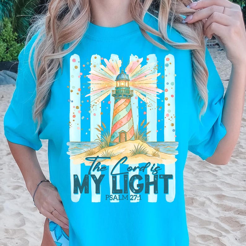Pu&ograve; includere: Maglietta turchese con un design a faro colorato. Il faro ha strisce rosse, arancioni e verdi. Il testo recita "The Lord is My Light" e Salmo 27:1. Il design include una scena di spiaggia con strisce blu e bianche.