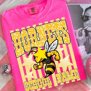 May include: A bright pink t-shirt with the word "HORNETS" in yellow block letters filled with white stars. A cartoon bee with yellow and black stripes is featured below the word. The words "SCHOOL NAME" are at the bottom.