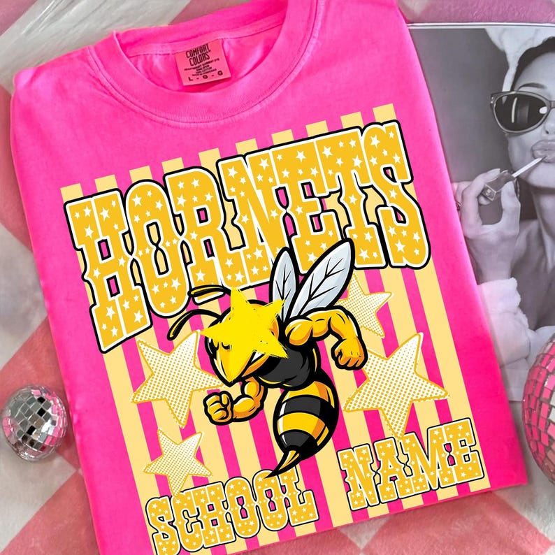May include: A bright pink t-shirt with the word "HORNETS" in yellow block letters filled with white stars. A cartoon bee with yellow and black stripes is featured below the word. The words "SCHOOL NAME" are at the bottom.