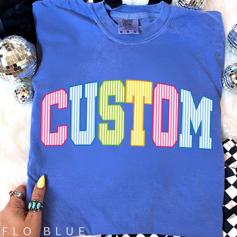 May include: A blue t-shirt with the word "CUSTOM" in large, colorful, striped letters. The letters are outlined in a contrasting color. The shirt has a crew neck and a small tag at the neckline. The text "FLO BLUE" is at the bottom.