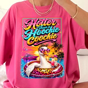May include: Pink t-shirt with a graphic featuring a goose wearing sunglasses and a leopard print headband, floating in a pool with a cocktail. The text reads "Hotter Than a Hoochie Coochie". Palm trees and a sunset are in the background.