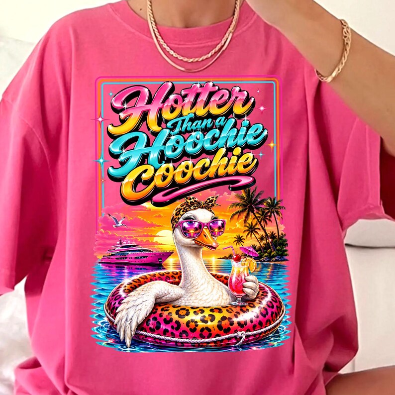 May include: Pink t-shirt with a graphic featuring a goose wearing sunglasses and a leopard print headband, floating in a pool with a cocktail. The text reads "Hotter Than a Hoochie Coochie". Palm trees and a sunset are in the background.