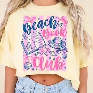 May include: A light yellow t-shirt with a graphic that reads "Beach Book Club." The design includes a book, seashells, a starfish, sunglasses, and a sun hat. The text "Members only dive into a good book" is also included.