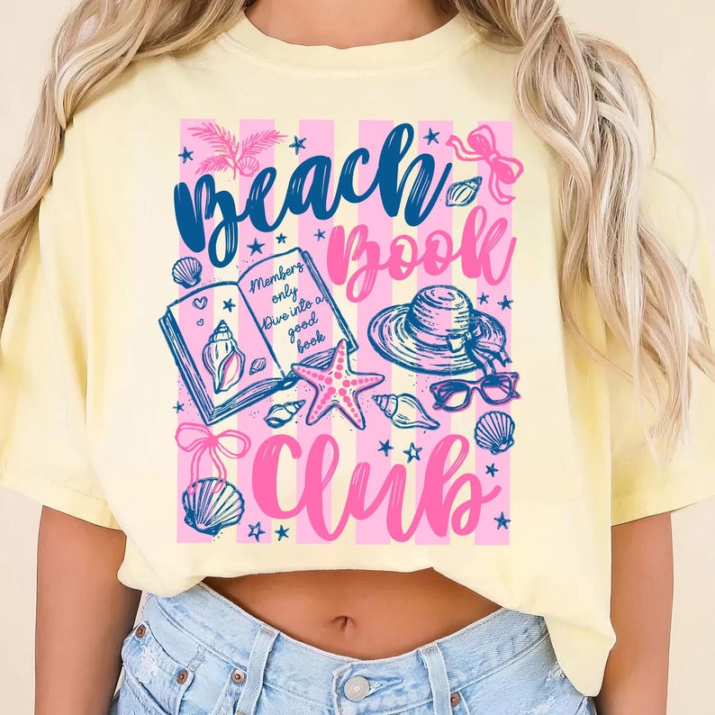May include: A light yellow t-shirt with a graphic that reads "Beach Book Club." The design includes a book, seashells, a starfish, sunglasses, and a sun hat. The text "Members only dive into a good book" is also included.