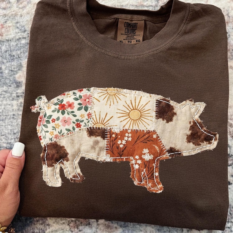 May include: A brown t-shirt with a patchwork pig design. The pig is made of various fabric patterns, including floral, sunburst, and cowhide prints. The shirt has a crew neck and a tag that says "Comfort Colors".