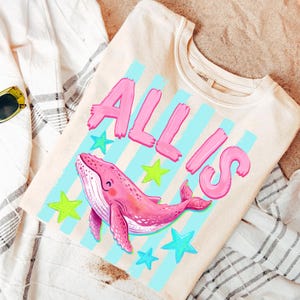 以下が含まれることがあります： ピンクのクジラのイラスト、ピンクの「ALL IS」の文字、青と緑の星が描かれたクリーム色のスウェットシャツ。 デザインは青い縦縞模様の上に配置されています。