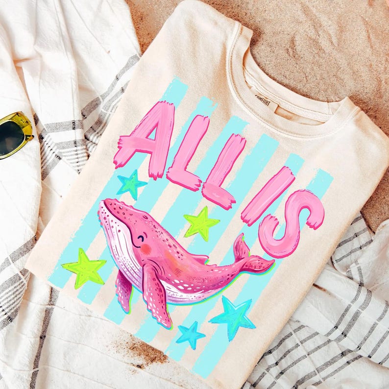 以下が含まれることがあります： ピンクのクジラのイラスト、ピンクの「ALL IS」の文字、青と緑の星が描かれたクリーム色のスウェットシャツ。 デザインは青い縦縞模様の上に配置されています。