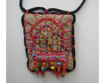 May include: A small, square, embroidered pendant with a colourful geometric design and a fringe of beads. The pendant is attached to a black cord.