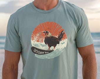 Könnte beinhalten: Hellblaues T-Shirt mit einem Surf-Hahn-Motiv. Der Hahn trägt eine Sonnenbrille und surft auf einer Welle, vor einem roten und orangefarbenen Sonnenuntergang. Der Text "LET'S CLUCKIN' RIDE!" ist auf dem Surfbrett aufgedruckt.
