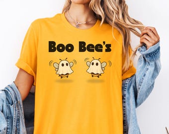 May include: Mustard yellow t-shirt with the text "Boo Bee's" in black. The shirt features two cartoon bees dressed as ghosts. The bees have white ghost costumes, black eyes, and are smiling. The shirt is a casual, everyday wear item.