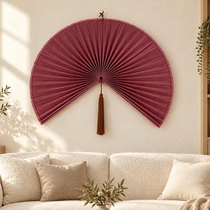 Peut inclure: Un grand éventail mural semi-circulaire de couleur bordeaux profond avec une texture plissée et un pompon marron foncé suspendu au centre. L'éventail est fixé au mur au-dessus d'un canapé de couleur claire, présentant un design symétrique.