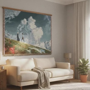 May include: A tapestry depicting a landscape with two figures walking on a hill, framed in wood. The artwork hangs above a cream-colored sofa with a blanket, a wooden side table, and a lamp. A potted plant sits nearby.