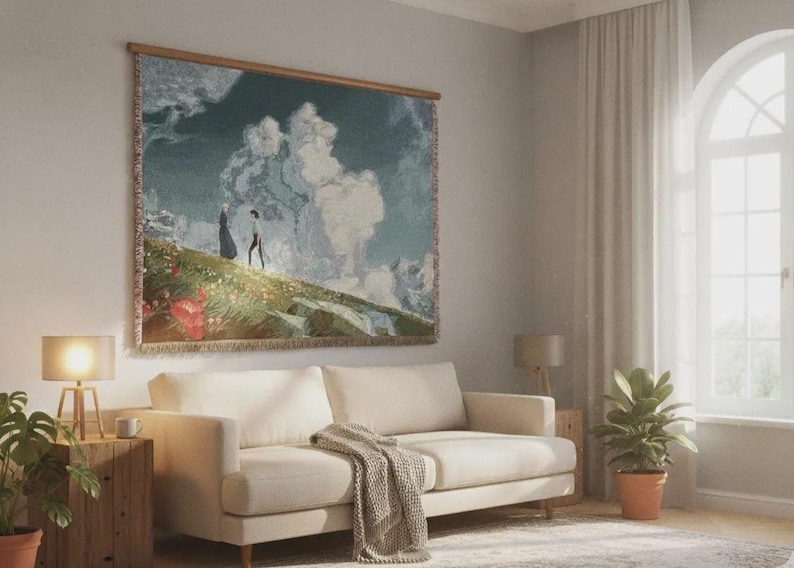 May include: A tapestry depicting a landscape with two figures walking on a hill, framed in wood. The artwork hangs above a cream-colored sofa with a blanket, a wooden side table, and a lamp. A potted plant sits nearby.