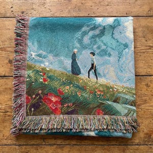 May include: A woven throw blanket featuring a scenic illustration of two figures on a grassy hill with red flowers, under a cloudy sky. The blanket has a fringed edge with shades of red, green, and blue.