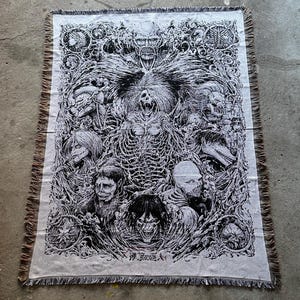 以下が含まれることがあります： 複数の人物と中心的な骨格の形を白黒で描いた、灰色の織りスローケット。 デザインには詳細なパターンとフリンジエッジが含まれており、下部に日本語のテキストがあります。
