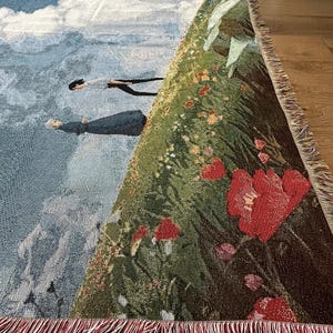 May include: Woven throw blanket featuring a scene with figures in the sky, clouds, and mountains. The blanket has a detailed illustration with red flowers and green foliage. The edges are finished with a red fringe.