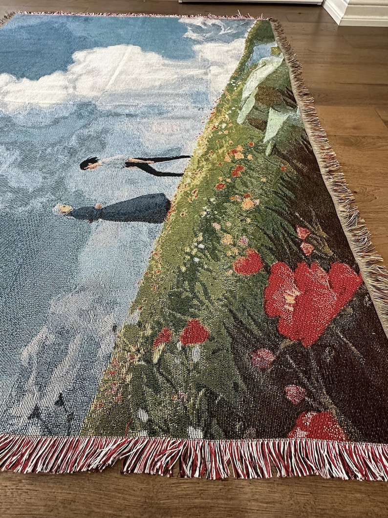May include: Woven throw blanket featuring a scene with figures in the sky, clouds, and mountains. The blanket has a detailed illustration with red flowers and green foliage. The edges are finished with a red fringe.