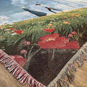 May include: Woven throw blanket featuring a pastoral scene with a field of red poppies, green grass, and a blue sky with clouds. The blanket has a fringed edge with red, white, and multicolored threads. The corner of the blanket is visible.