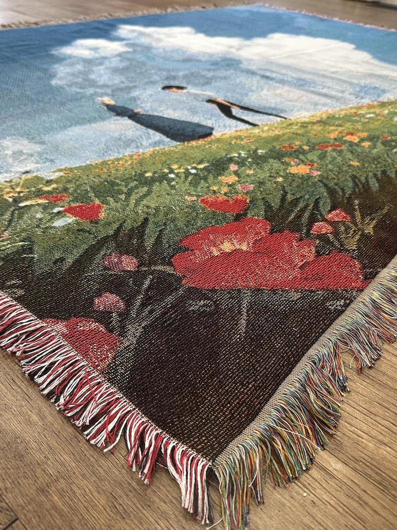 May include: Woven throw blanket featuring a pastoral scene with a field of red poppies, green grass, and a blue sky with clouds. The blanket has a fringed edge with red, white, and multicolored threads. The corner of the blanket is visible.