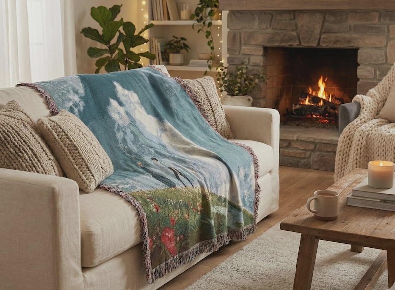 May include: A woven throw blanket with a landscape design in shades of blue, green, and white is draped over a beige sofa. The blanket features a fringed edge and is paired with knitted throw pillows. A lit fireplace is in the background.