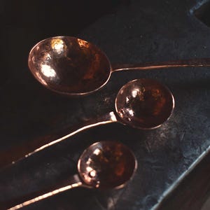 May include: Three copper spoons of varying sizes arranged in a diagonal line. Each spoon features a round, hammered bowl and a long, slender handle. The metal has a warm, reddish-brown metallic finish with visible indentations from the hammering.