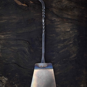 May include: Hand-forged metal spatula with a twisted handle and a looped end. The spatula head is rectangular with rounded corners and is attached to the handle with two rivets. The metal has a silver and blue iridescent finish.