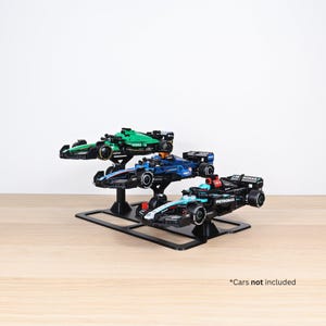 Op de afbeelding: Een zwarte displaystandaard met drie speelgoed raceauto's. De auto's zijn groen, blauw en zwart met sponsorlogo's. De standaard staat op een licht houten oppervlak. De tekst onderaan luidt "*Auto's niet inbegrepen."