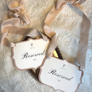 May include: Two white "Reserved" signs with a pink border and elegant script, tied with blush-colored ribbons. The signs are placed on a decorative mirror, set against a textured white background. The signs have the date "October 15, 2023".