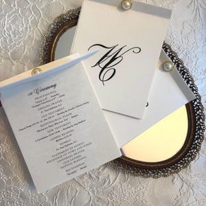 May include: Elegant wedding ceremony programs and stationery. The programs are white with black text and a gold pearl accent. The programs are displayed on a decorative mirror with a gold frame.