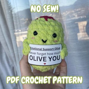 Könnte beinhalten: Eine handgefertigte, hellgrüne Häkelolive mit einem lächelnden Gesicht, schwarzen Augen und einem roten Oberteil. Sie hat ein weißes Etikett mit der Aufschrift "Emotional Support Olive" und "OLIVE YOU". Der Text "NO SEW!" und "PDF CROCHET PATTERN" sind ebenfalls sichtbar.