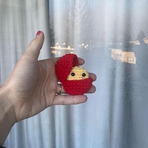 May include: A small crocheted toy in the shape of a person with a red crocheted shell. The toy has a yellow face with black eyes and a small smile.