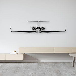 Puede incluir: Una pieza de arte mural de metal negro con la forma de la vista frontal de un avión jet privado. El diseño presenta una silueta simétrica con alas largas y delgadas, dos tomas de aire circulares y una configuración de cola en T, en estilo lineal.