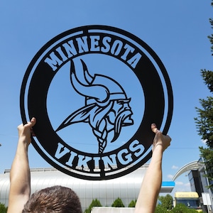 Könnte beinhalten: Ein schwarzes Metallschild mit den Worten "Minnesota Vikings" in weißen Buchstaben. Das Schild zeigt ein stilisiertes Bild eines Wikingerhelms mit Hörnern.