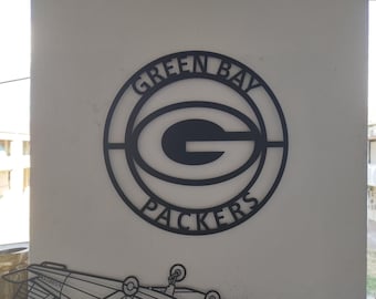 Puede incluir: Un letrero de pared circular de metal negro con el logotipo de los Green Bay Packers en el centro. El texto GREEN BAY está arqueado a lo largo del círculo interior superior, y la palabra PACKERS está arqueada a lo largo del círculo inferior, rodeando la G.