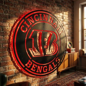 May include: A circular Cincinnati Bengals wall sign featuring a black metal frame with a cut-out logo and text. The sign is illuminated from behind with a bright red light, highlighting the team name and the stylized B logo against the brick wall.