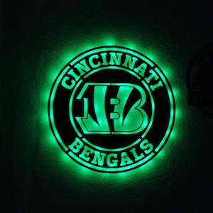 May include: A circular wall sign featuring the Cincinnati Bengals logo and text illuminated by a bright green LED light. The design consists of a black circular frame with the team name and the stylized B logo cut out, allowing the green light to shine through.